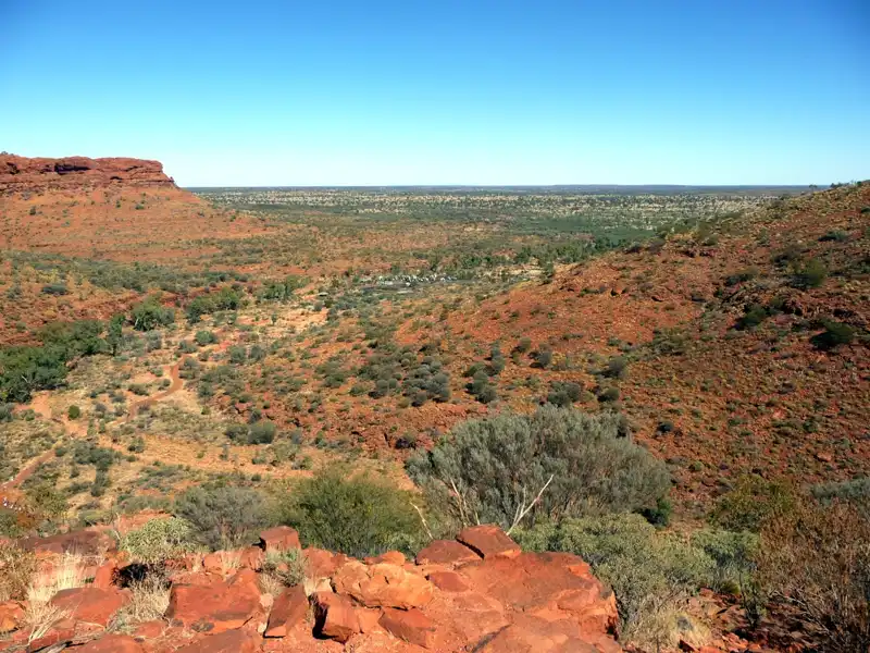 A wide-angle panoramic view of the Australian outback featuring vibrant red soil, sparse green shrubs, and a clear blue horizon stretching into the distance.