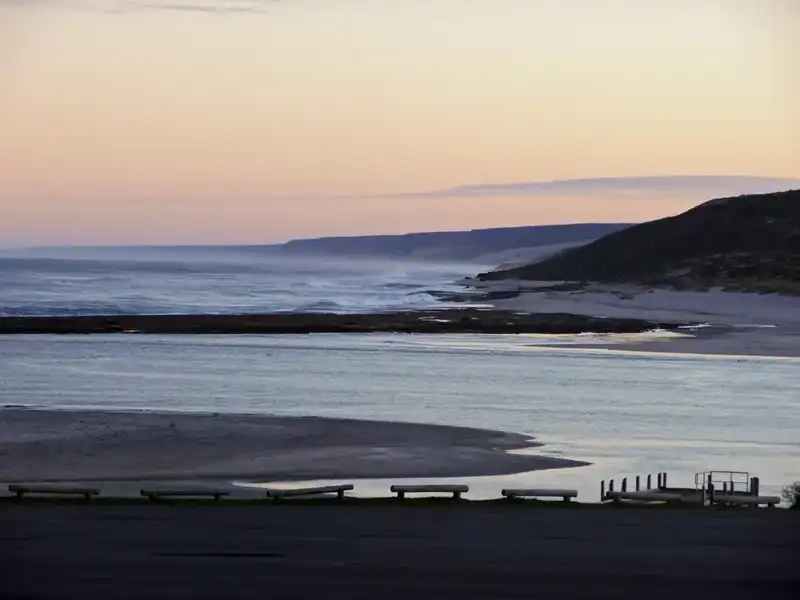 A coastal landscape at sunset shows calm waters meeting the sandy shore, with gentle waves and distant cliffs under a pastel-colored sky. A small jetty and barriers are visible in the foreground, reminiscent of scenes along the Ningaloo Coast.