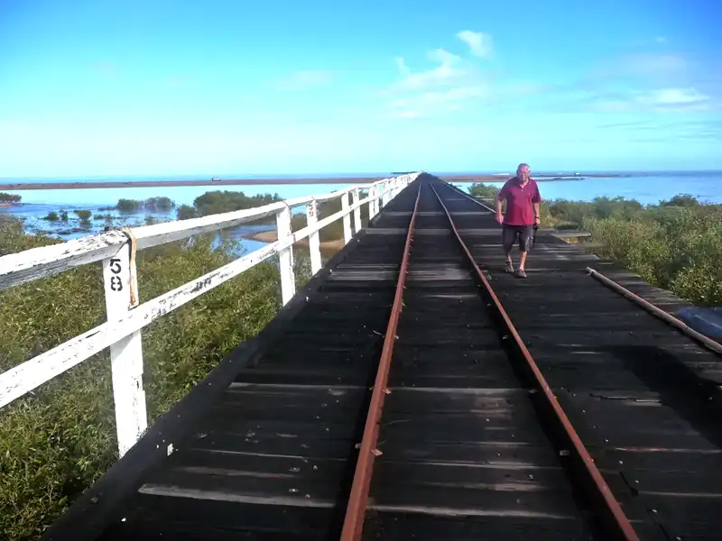A person walks on an old wooden railway bridge with white railings, surrounded by water and greenery under a bright blue sky—an idyllic scene reminiscent of the landscapes found on an Exmouth to Perth road trip. The bridge extends into the distance.
