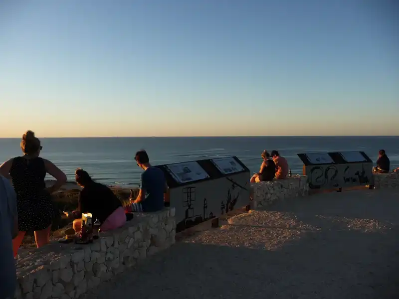 People sit and stand near informational signs on a stone lookout, facing the ocean during sunset, with clear skies and calm water in the background—an ideal stop on an Exmouth to Perth road trip exploring sights like Shark Bay stromatolites.