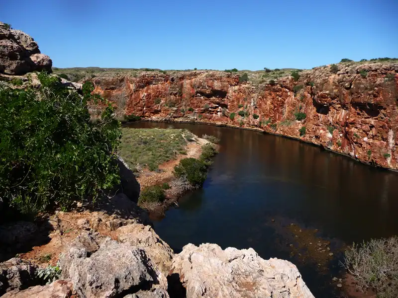 Rocky, reddish cliffs curve around a calm, dark river beneath the clear blue sky of the Ningaloo Coast, with green shrubbery growing along the landscape and some vegetation near the water’s edge.