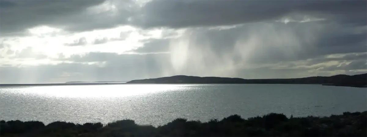 A large, calm body of water reflects bright sunlight under a cloudy sky, with distant low hills and streaks of rain falling beyond. Dark vegetation lines the shore in the foreground, evoking scenes along the Ningaloo Coast on an Exmouth to Perth road trip.