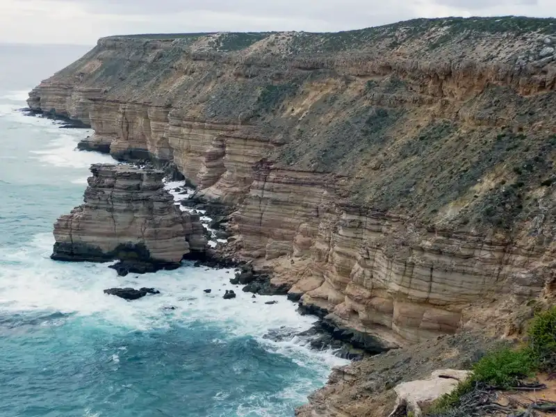 Steep, layered sandstone cliffs rise above turquoise ocean waves near rocky outcrops and sparse vegetation—an iconic sight along the Exmouth to Perth road trip under a cloudy sky.