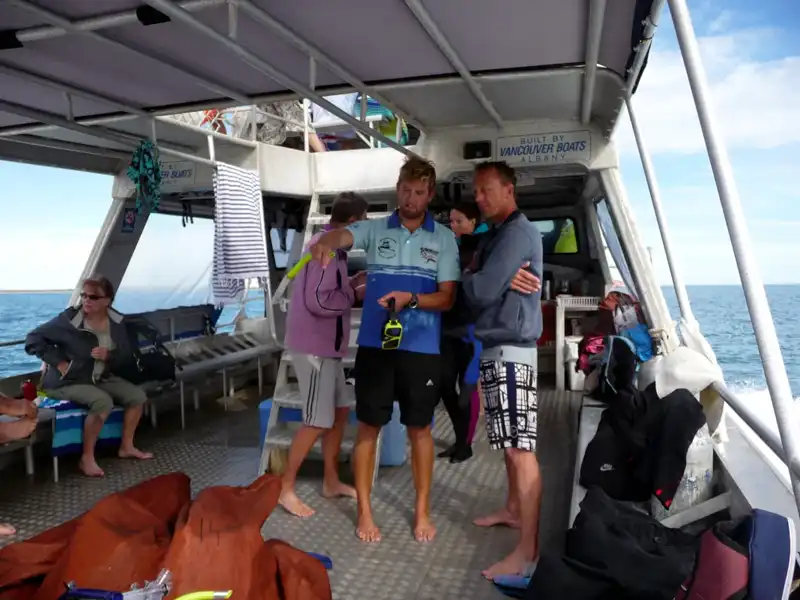 A group of people stand and sit on a boat deck, some holding snorkeling gear, ready to explore the manta rays Coral Bay is known for. The boat drifts at sea under a partly cloudy sky, bags and towels scattered around as excitement builds.
