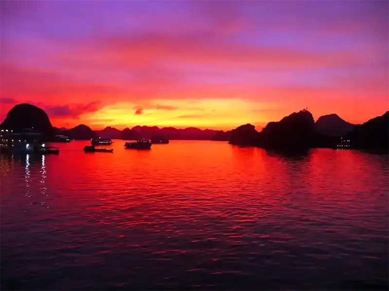 Sunset over Ha Long Bay with limestone karst islands silhouetted against a vibrant orange and red sky reflected in calm emerald water, Vietnam.