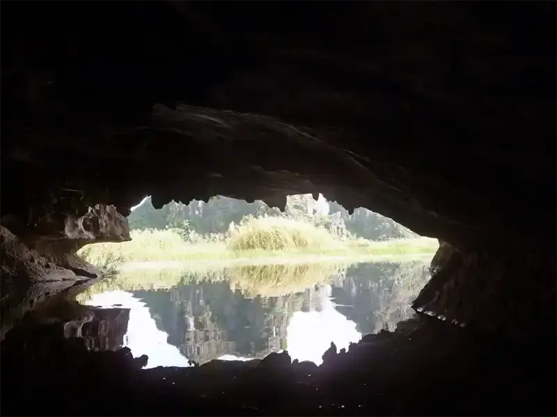 Boat entering a dark, low-ceilinged cave with still water reflecting the cave walls like a mirror.